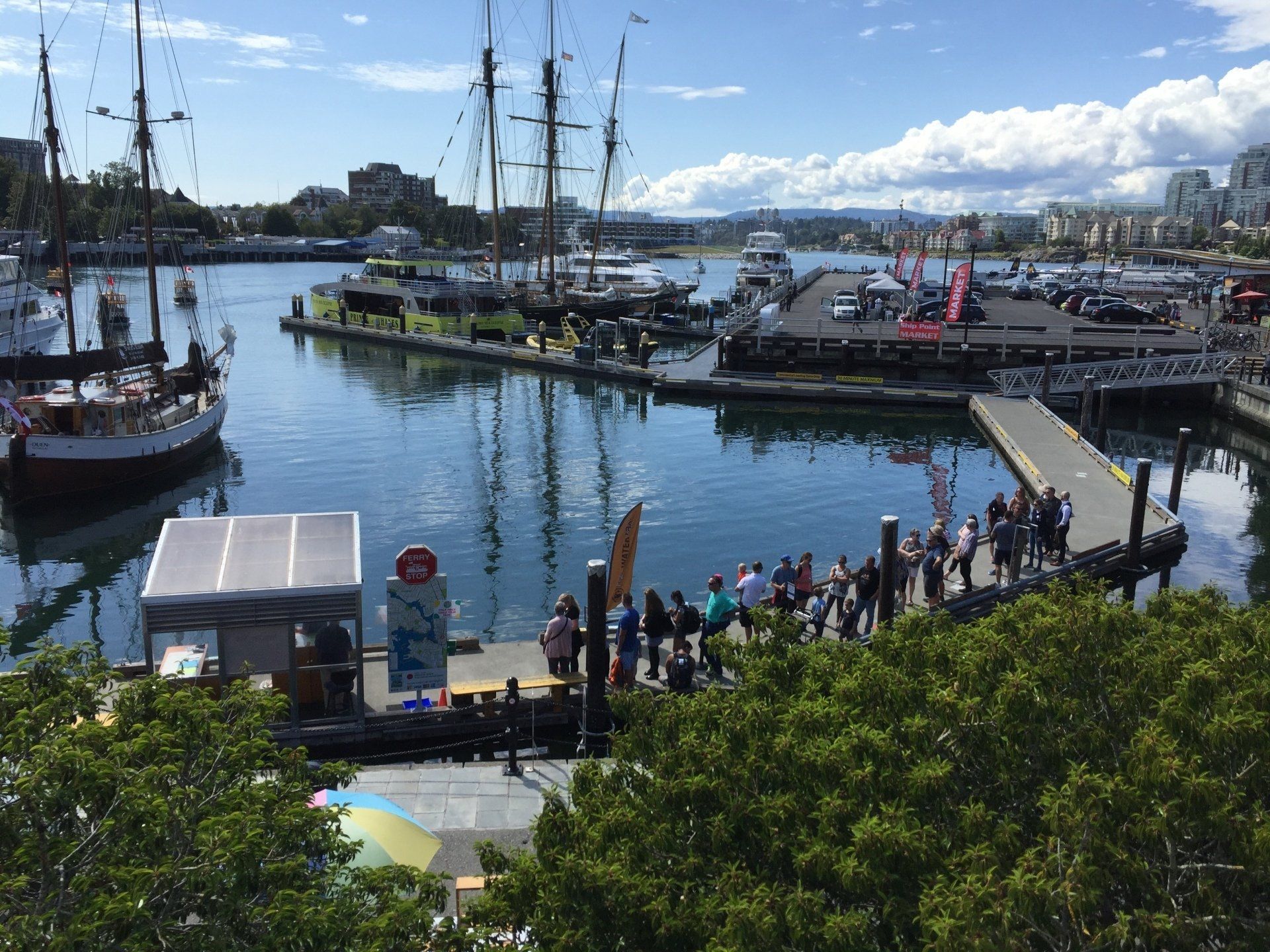 Harbor with sailboats and a crowd gathered on a pier; sunny day with blue sky and water.
