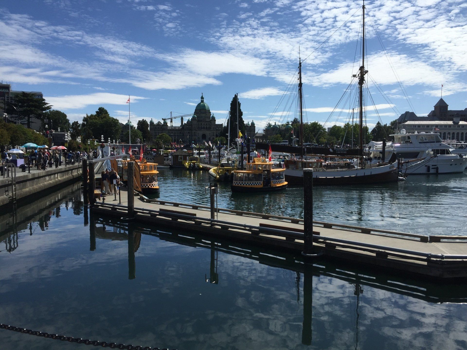 Boats docked in a harbor; city skyline in the background; people walking along the boardwalk.