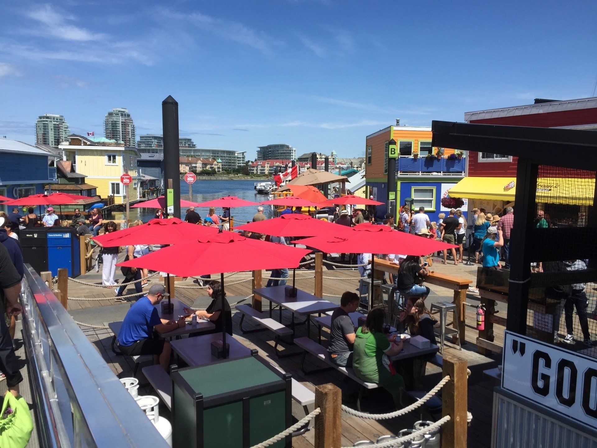 Outdoor restaurant with red umbrellas, people, and colorful buildings. Waterfront setting on a sunny day.