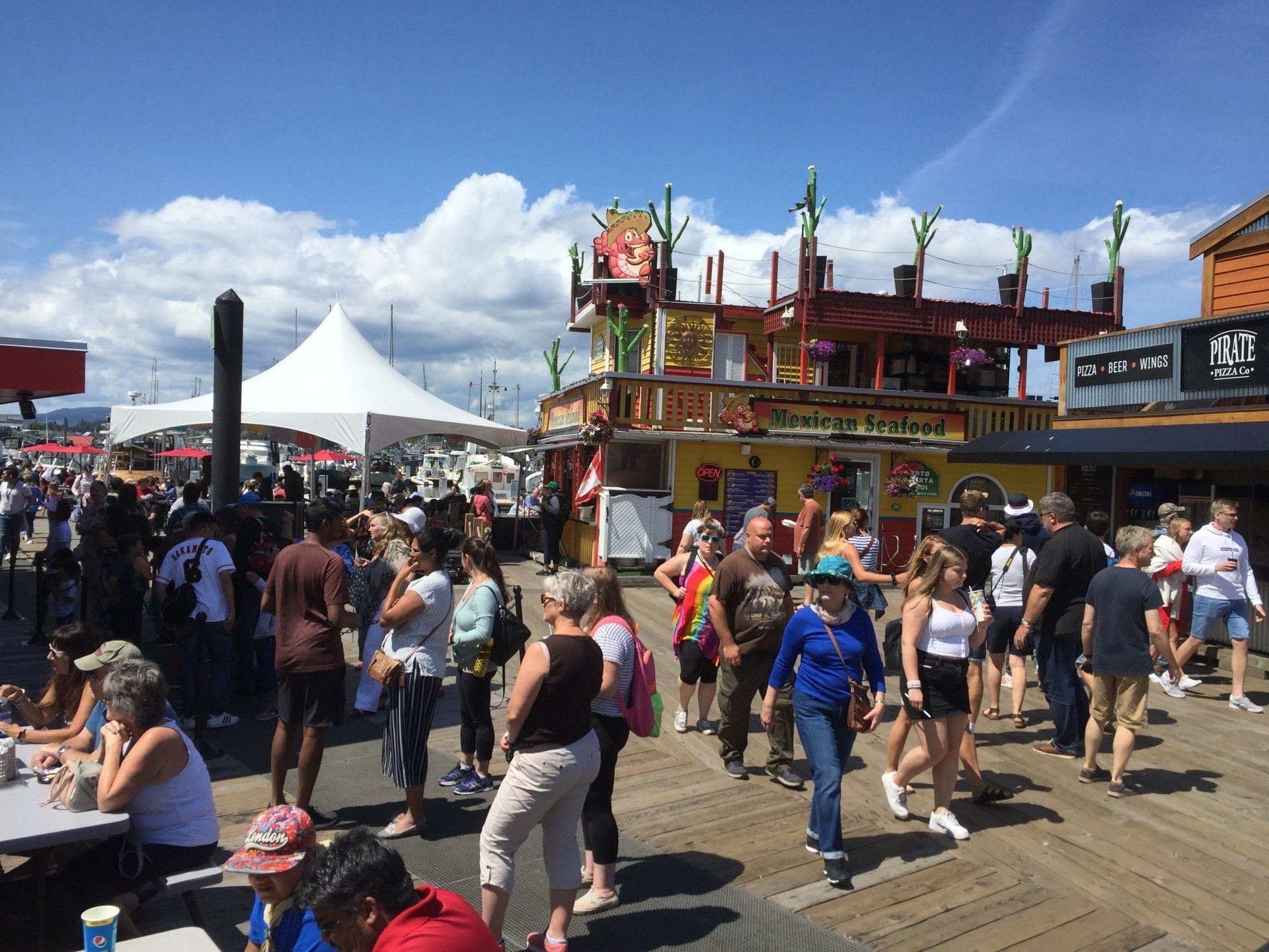 Crowd on boardwalk near food vendors under a sunny sky; people eating, walking.