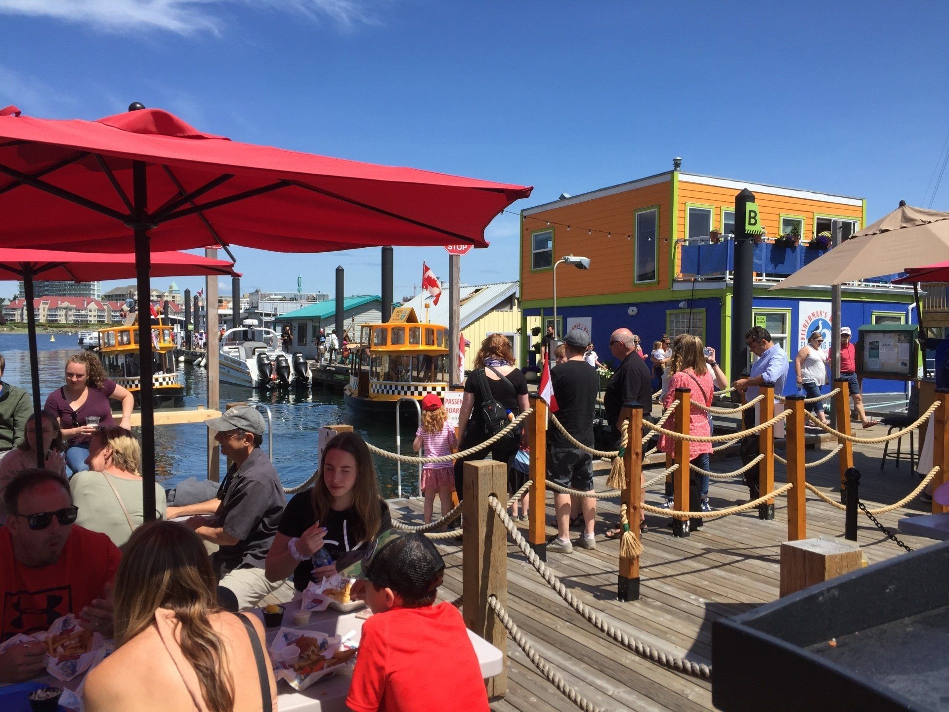 People dining at a waterfront restaurant with red umbrellas and colorful buildings in the background.