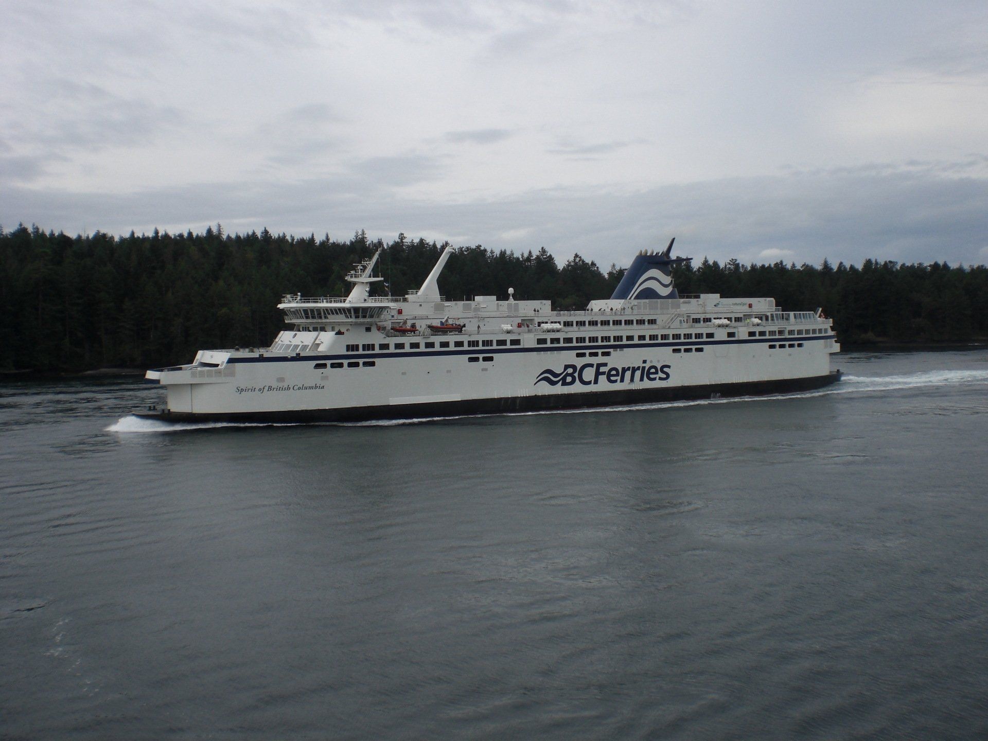 A white BC Ferries vessel sails on calm water near a wooded shoreline under a cloudy sky.