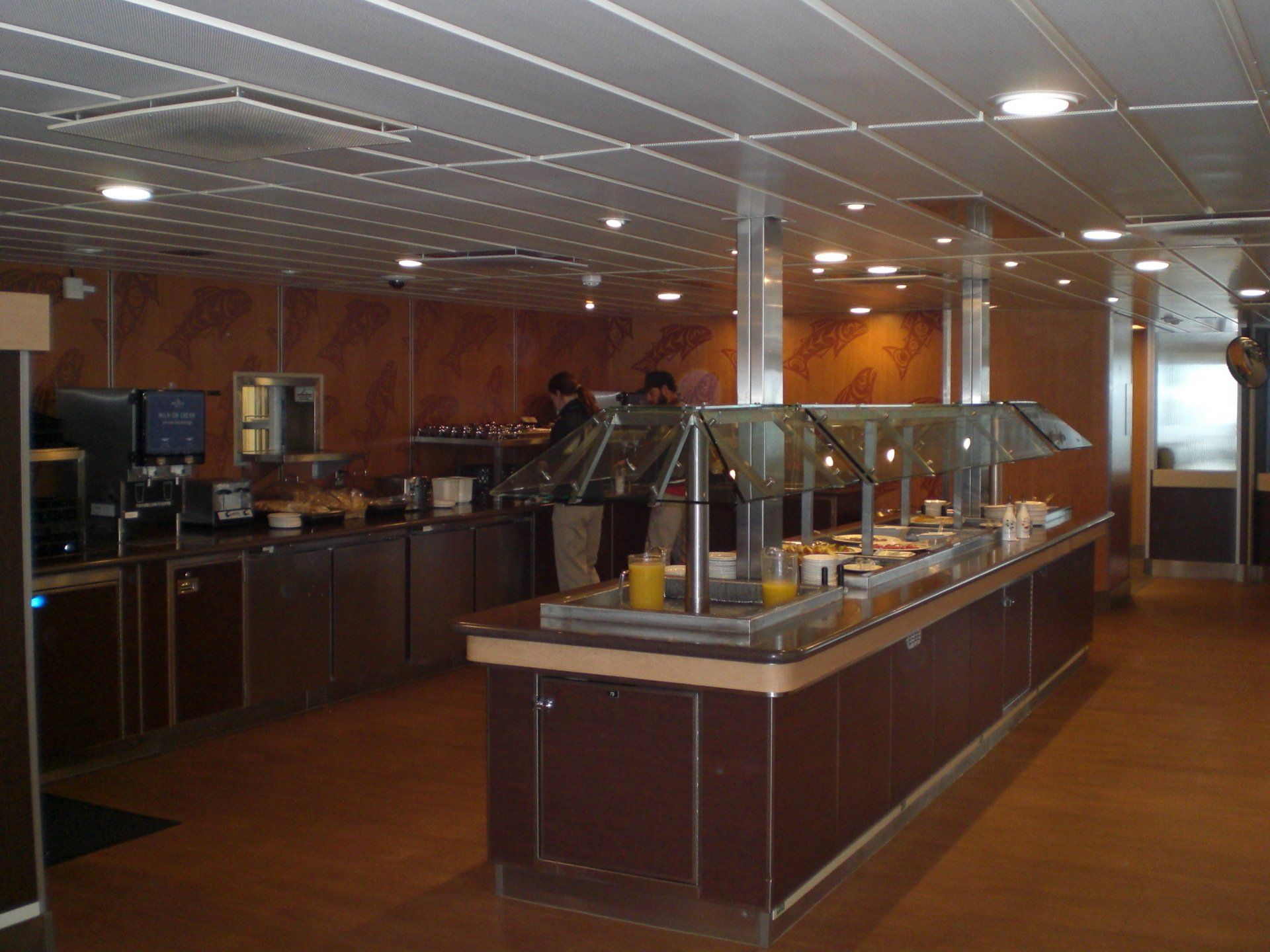 Buffet area with brown counters, stainless steel accents, and food under glass, possibly on a cruise ship.