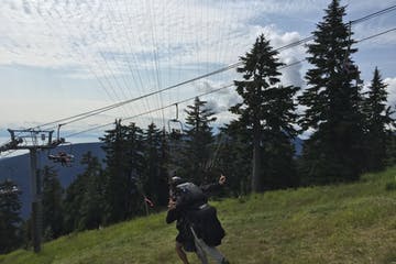 Person with parachute preparing to launch from a grassy mountain slope, chairlift in the background.