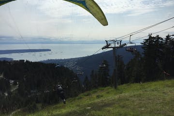 Paraglider soaring above a forested hillside, overlooking a city and the ocean.