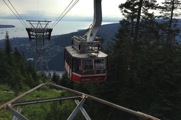 Cable car descending a mountain, red and white car with passengers. Coastal city and water in the background.