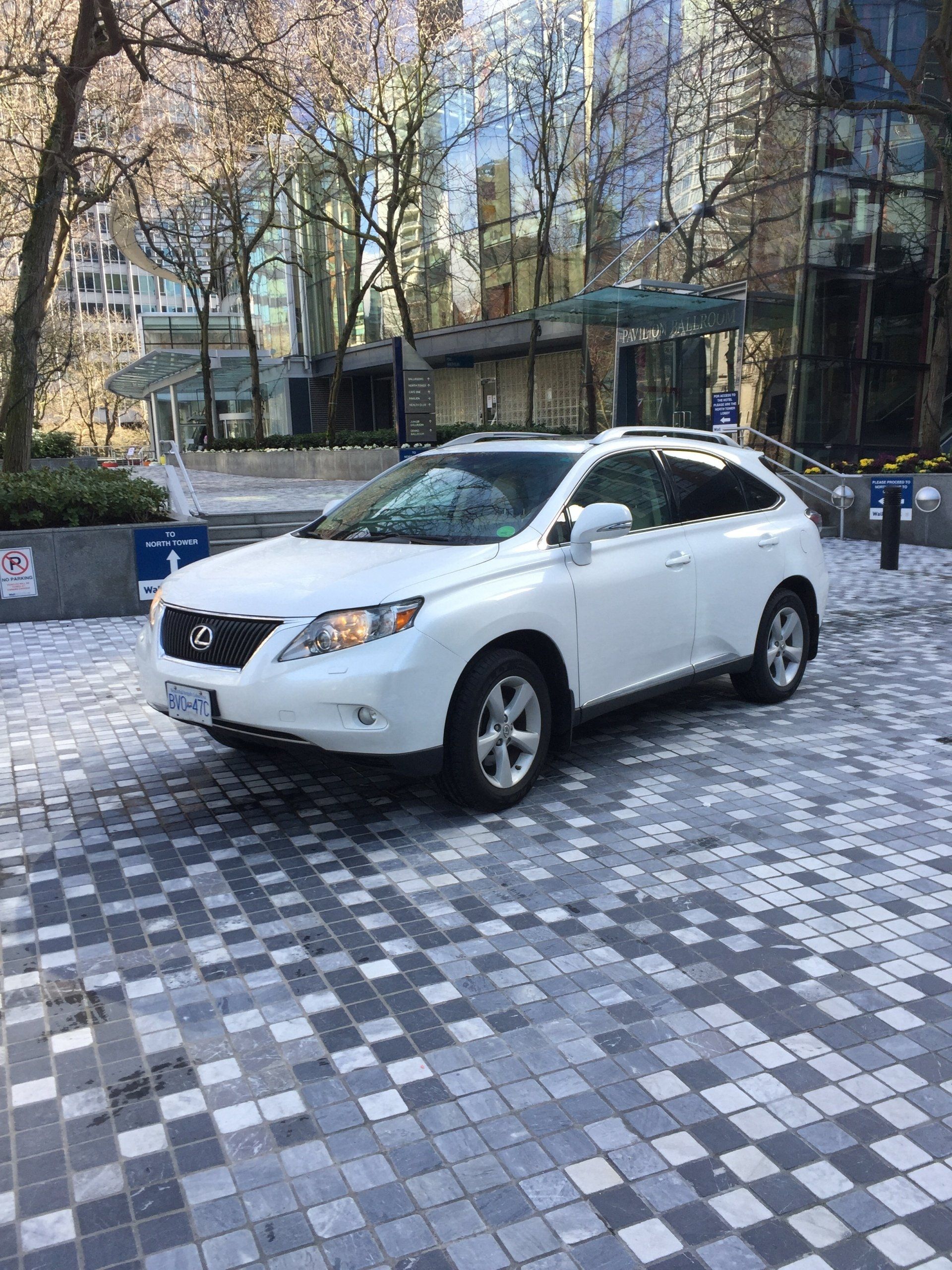 White Lexus SUV parked on a brick-paved plaza in front of a modern building with trees.