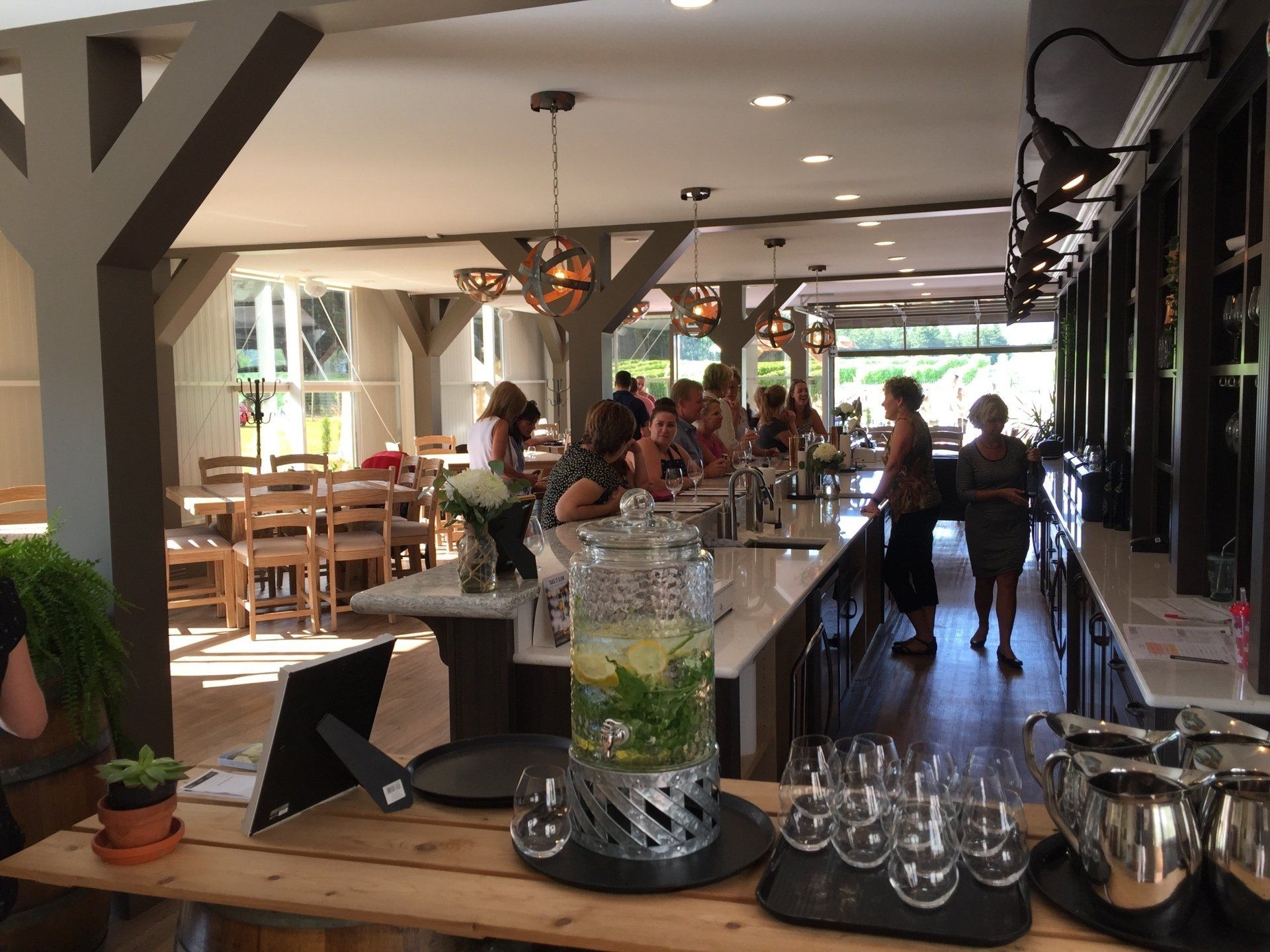 Bright bar with patrons; refreshments, a long counter, and light wooden beams.