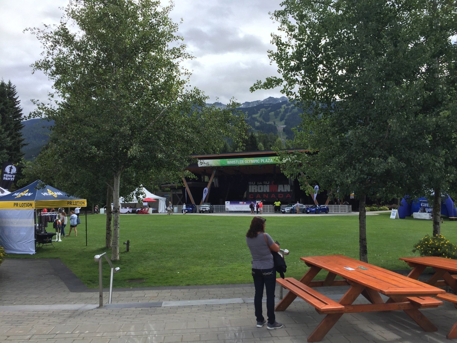 Park with a stage, picnic tables, and trees; a woman stands in front.