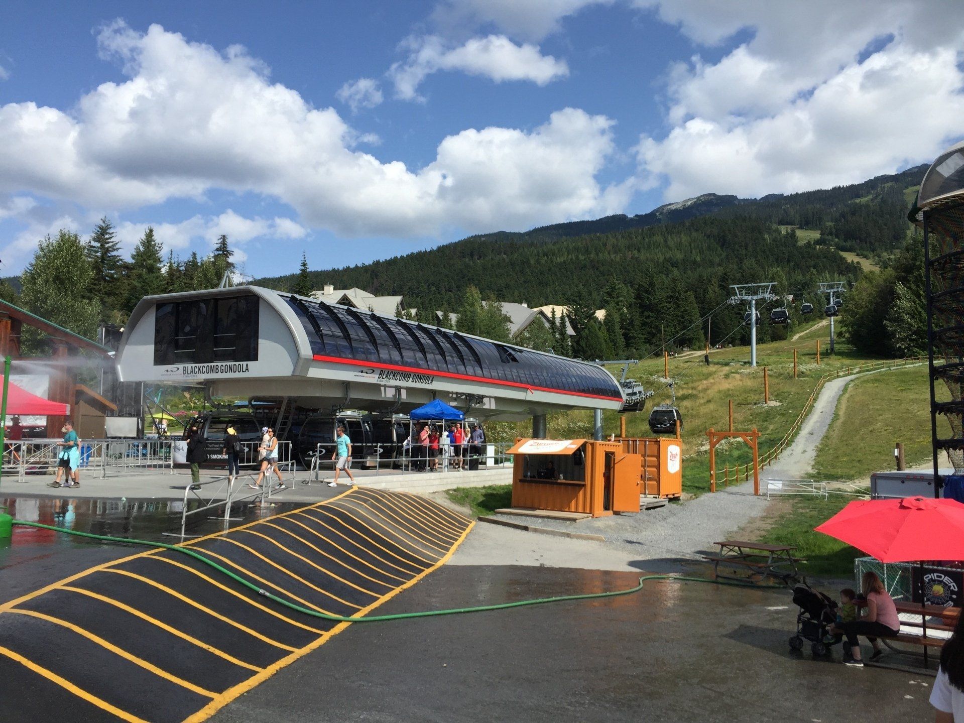 A ski lift station in summer with people, mountains, and a bright sunny sky.