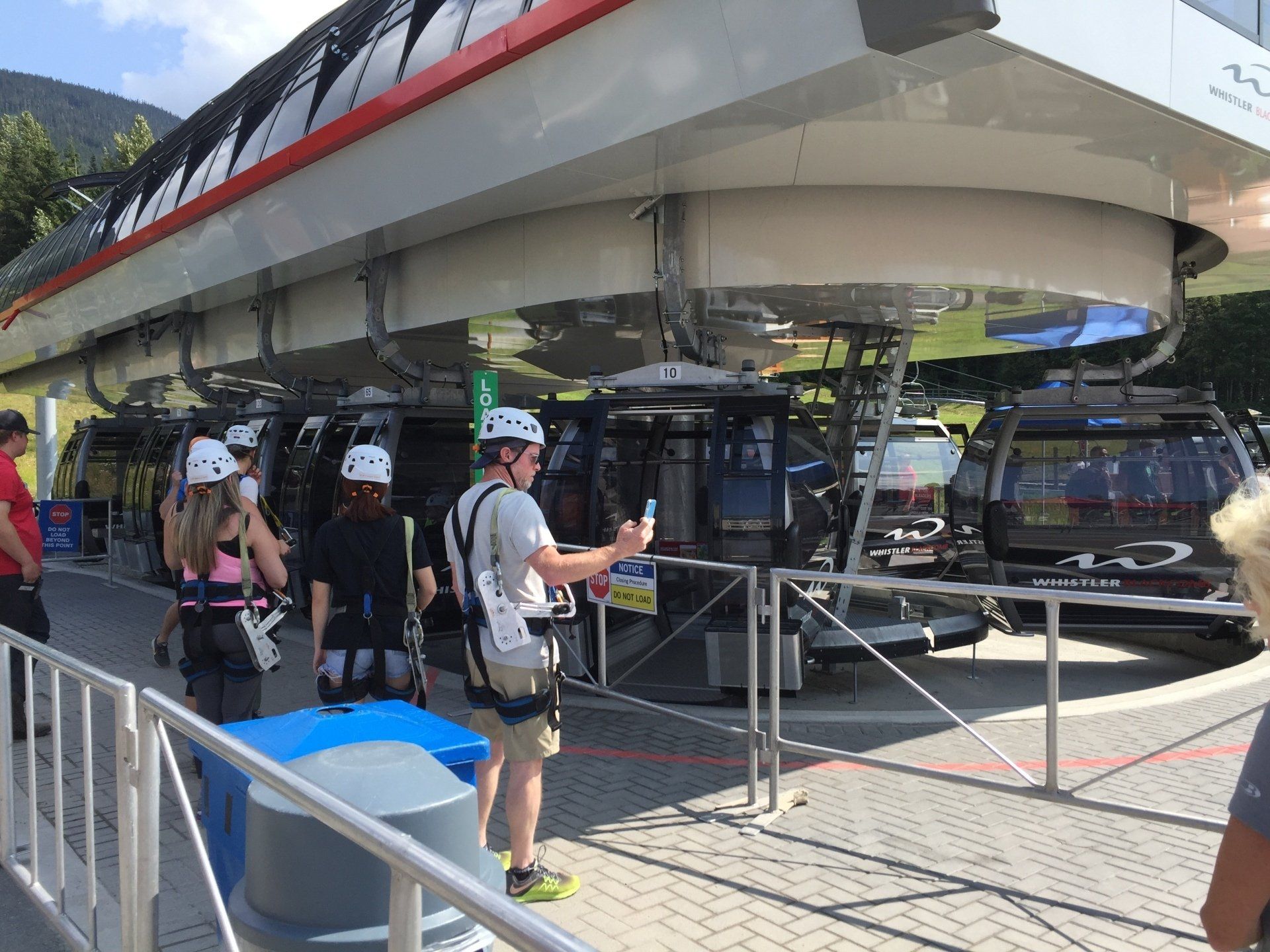 People in harnesses by gondola station, taking selfies, in a sunny outdoor setting.