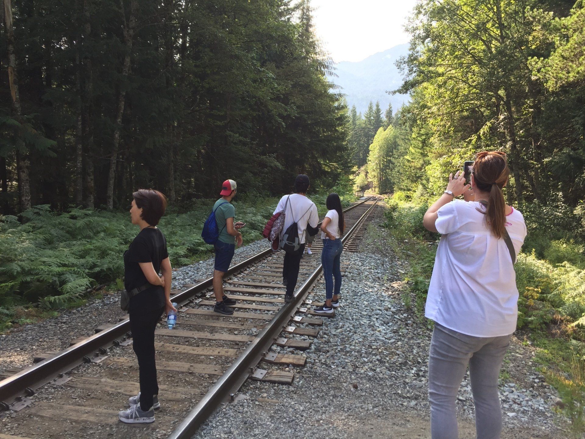 People walking on railroad tracks in a forest, one taking a photo.