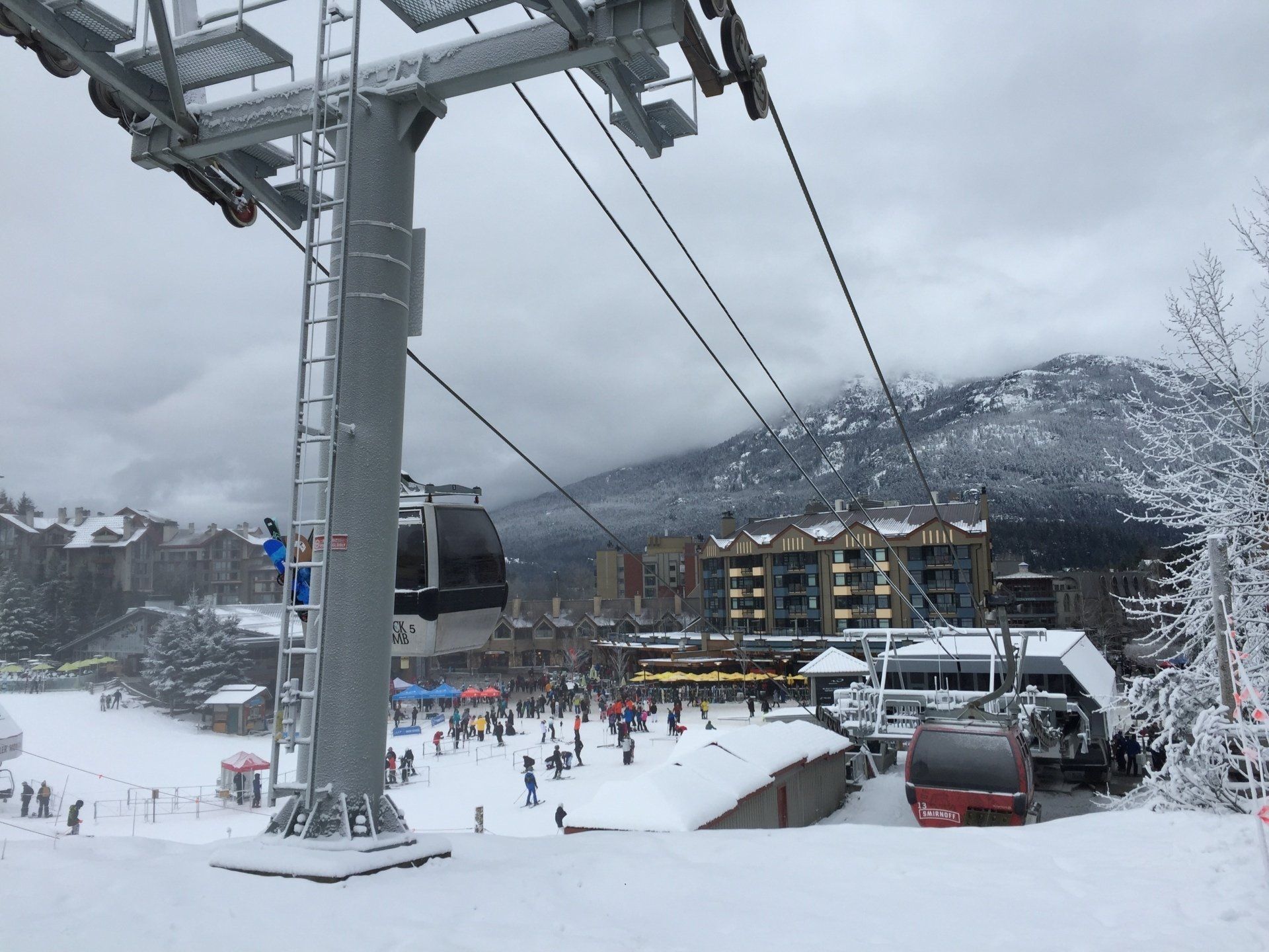 Ski lift at a snowy resort with people skiing, with buildings and mountains in the background.