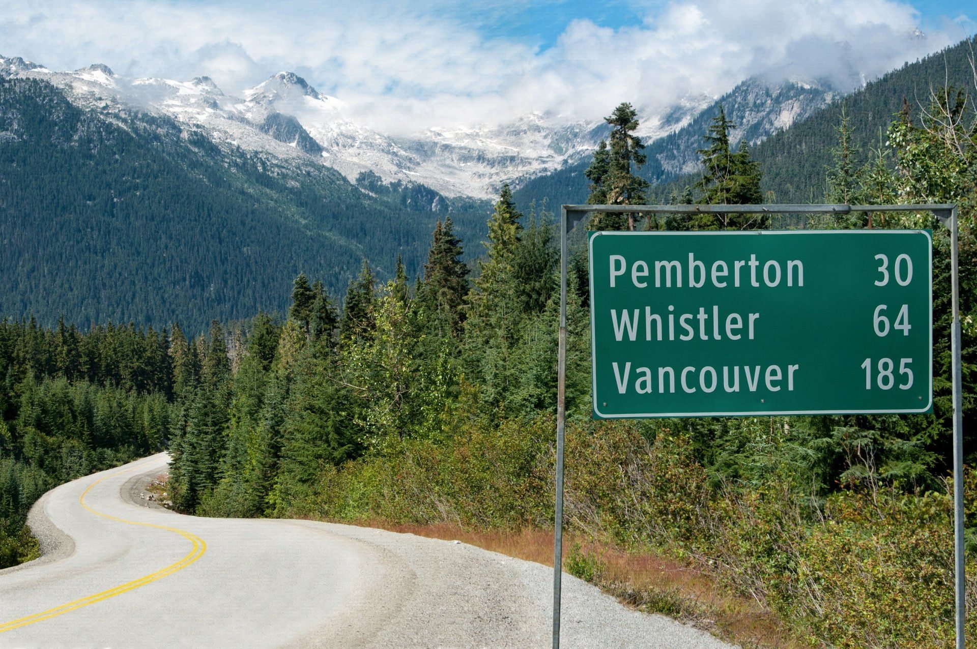 Road sign: Pemberton 30 km, Whistler 64 km, Vancouver 185 km; road through mountains, forest, and snowy peaks.