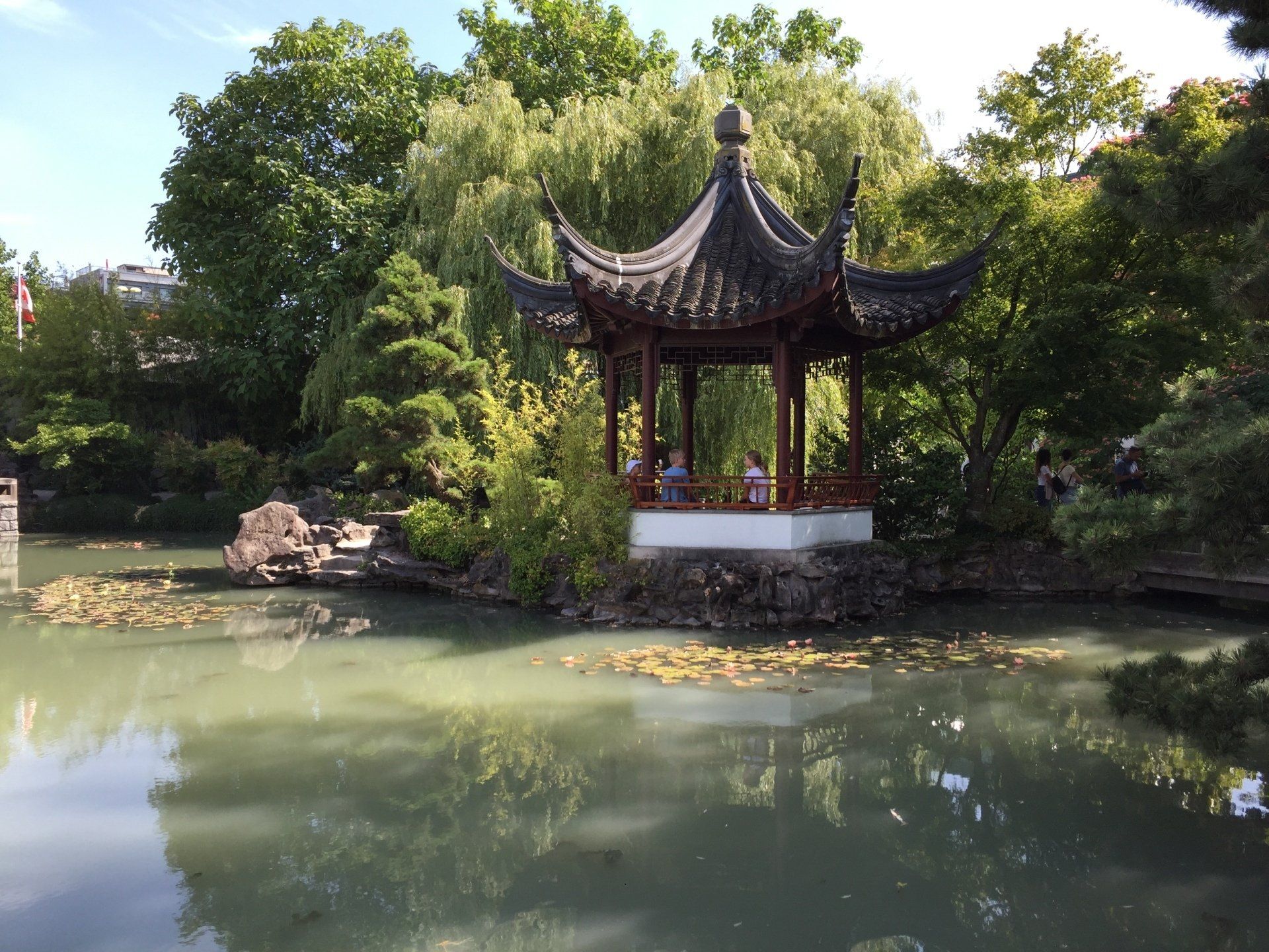 Chinese pagoda on a pond, surrounded by greenery; two people visible inside.