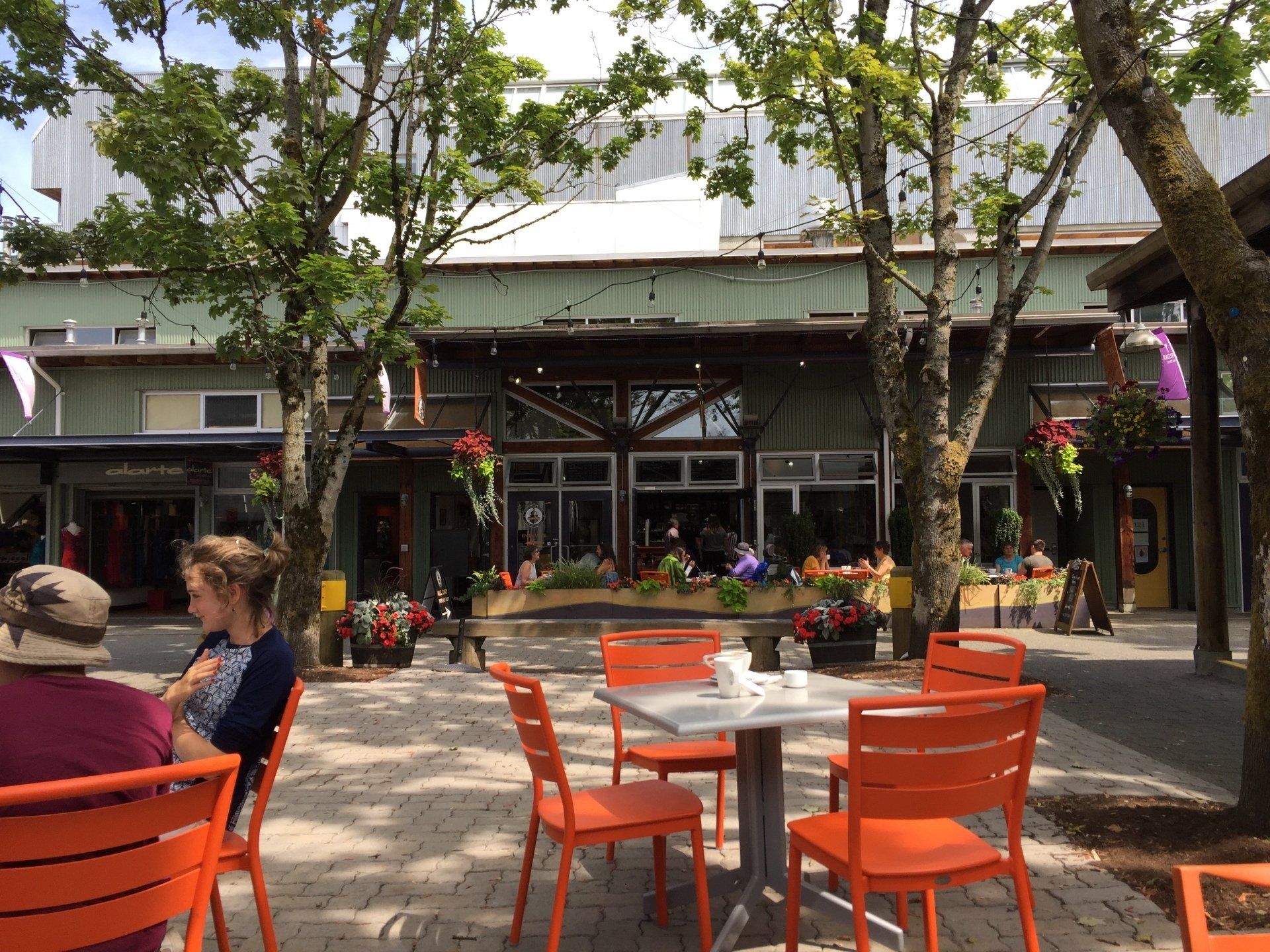 Outdoor cafe with orange chairs, people, and building with large windows and flowers.