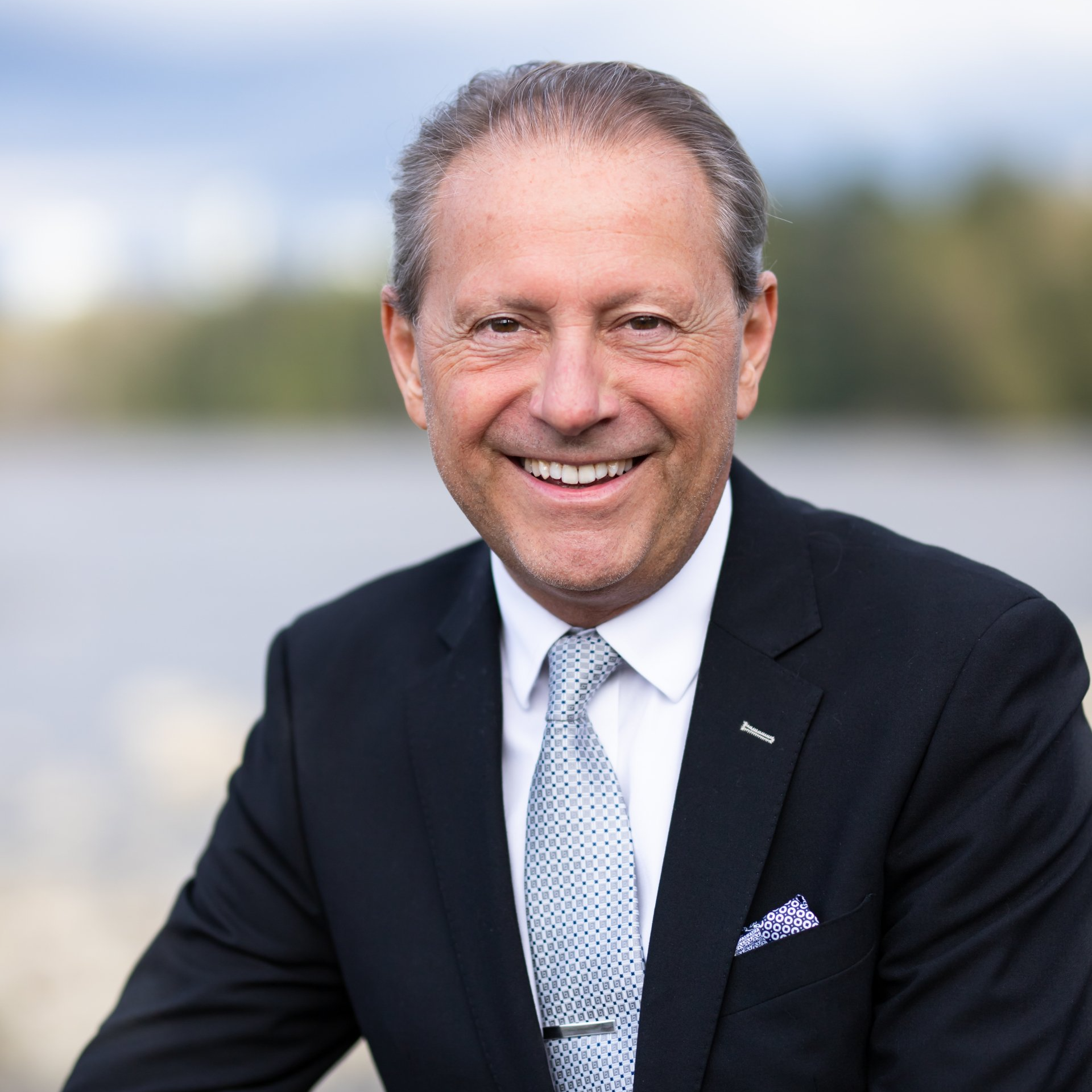 Smiling man in a suit, light blue tie, overlooking a lake with trees in the background.