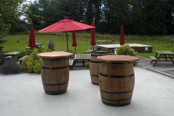 Three wooden barrel tables with red umbrellas on a patio with picnic tables in the background at Fraser Valley vineyards
