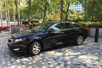 Black Kia sedan parked on a paved area, trees and a building in the background.