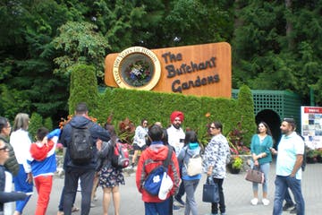 People at The Butchart Gardens entrance; wooden sign, greenery, tourists, sunny day.