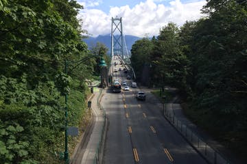 Lions Gate Bridge in Vancouver, BC. A car-filled road leads to the bridge, flanked by trees.
