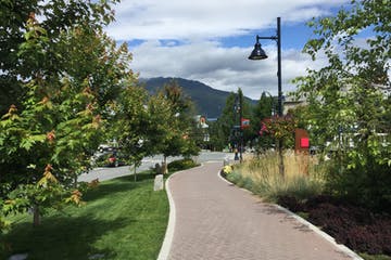 Paved path winds through a park-like setting. Trees frame the path, leading to a mountain view under a cloudy sky.