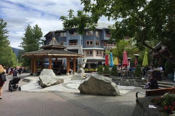 A plaza with a wooden gazebo, large rocks, and outdoor restaurant seating in front of a blue building, Whistler, BC.