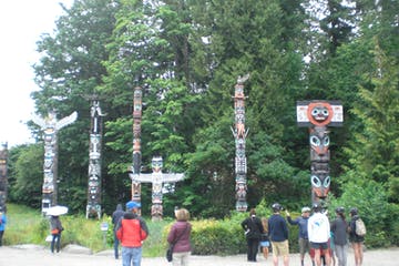 Totem poles standing in a park with people viewing. Green trees and overcast sky in background.