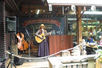 Musician on stage at Capilano Suspension Bridge, playing guitar. Wooden stage, cello, crowd in background.