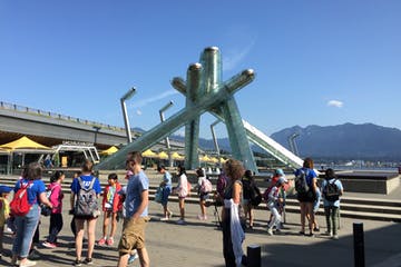 People gather near the Olympic Cauldron sculpture in Vancouver, with mountains in the background on a sunny day.