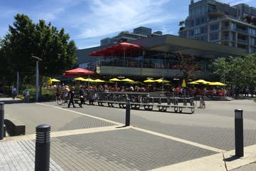 Outdoor restaurant with red and yellow umbrellas. People seated at tables. Buildings in the background.