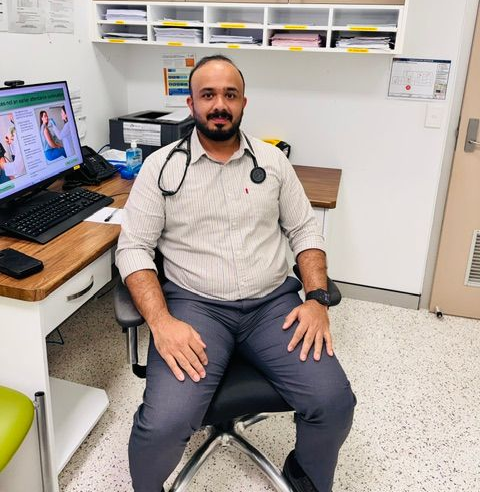 Doctor seated in a clinic exam room with a stethoscope, computer monitor, and medical supplies nearby