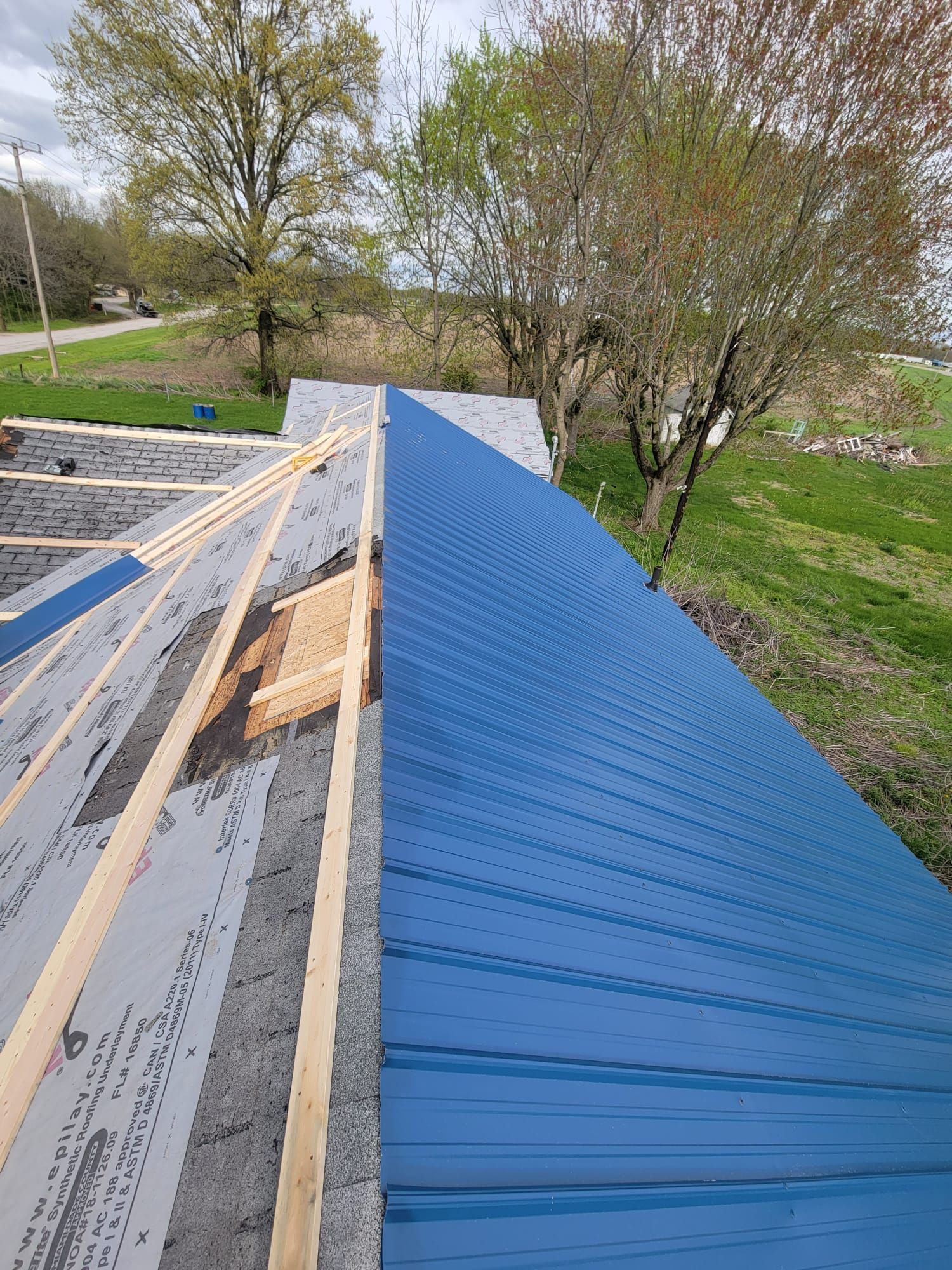 Blue metal roof under construction beside exposed framing and trees in a grassy yard