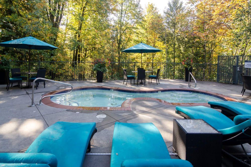 Two hot tubs, blue lounge chairs, and tables with umbrellas surrounded by trees at Sofi at Forest Heights, offers apartments for rent in Portland, OR.
