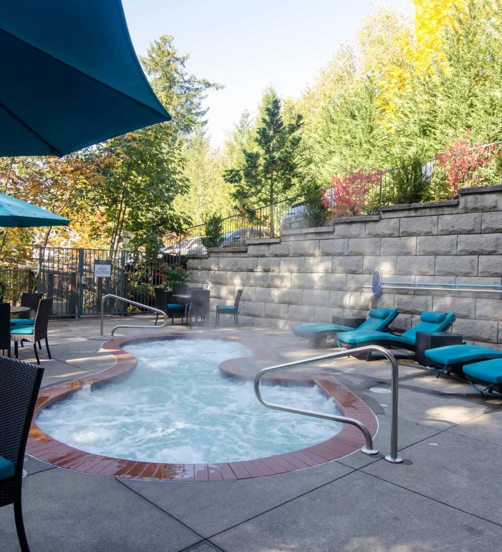 Outdoor hot tub with turquoise lounge chairs, umbrellas, and stone retaining wall, fall foliage in the background at Sofi at Forest Heights, offers apartments for rent in Portland, OR.