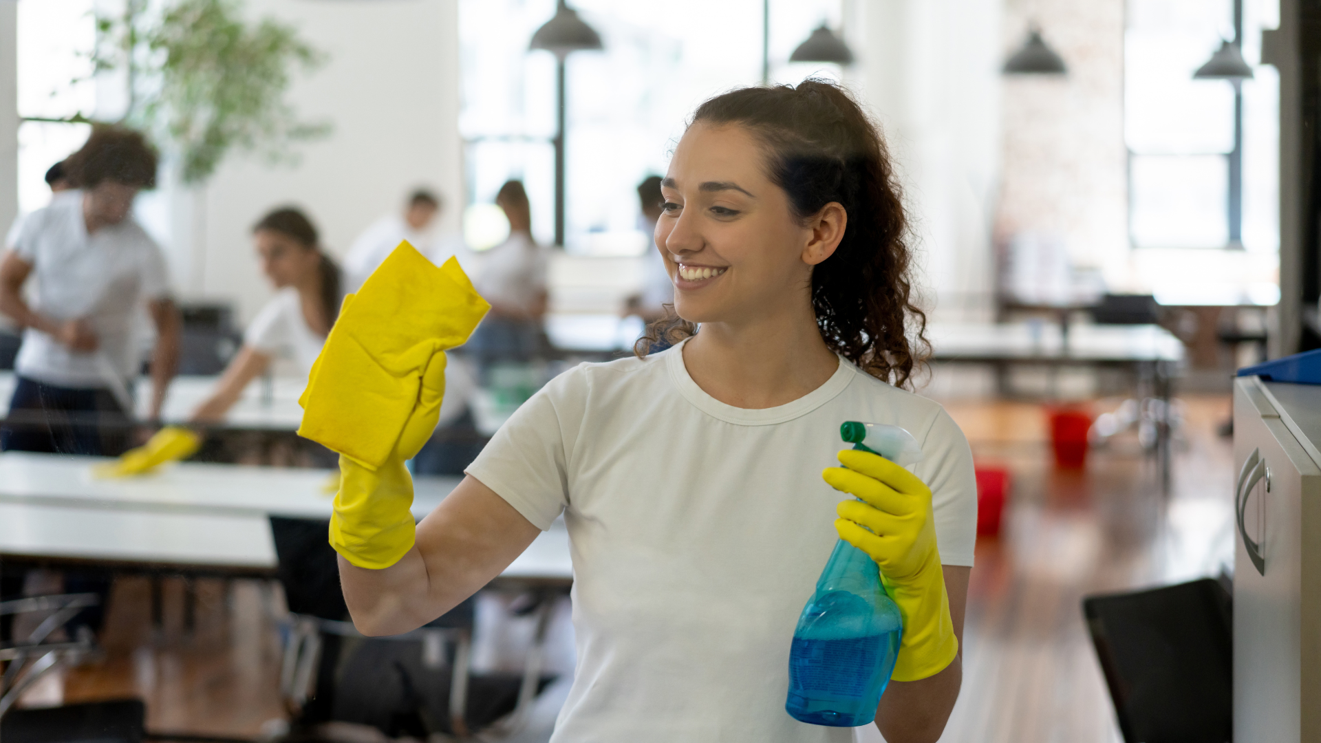 Person in yellow gloves and blue spray bottle cleaning a table in a bright restaurant, smiling