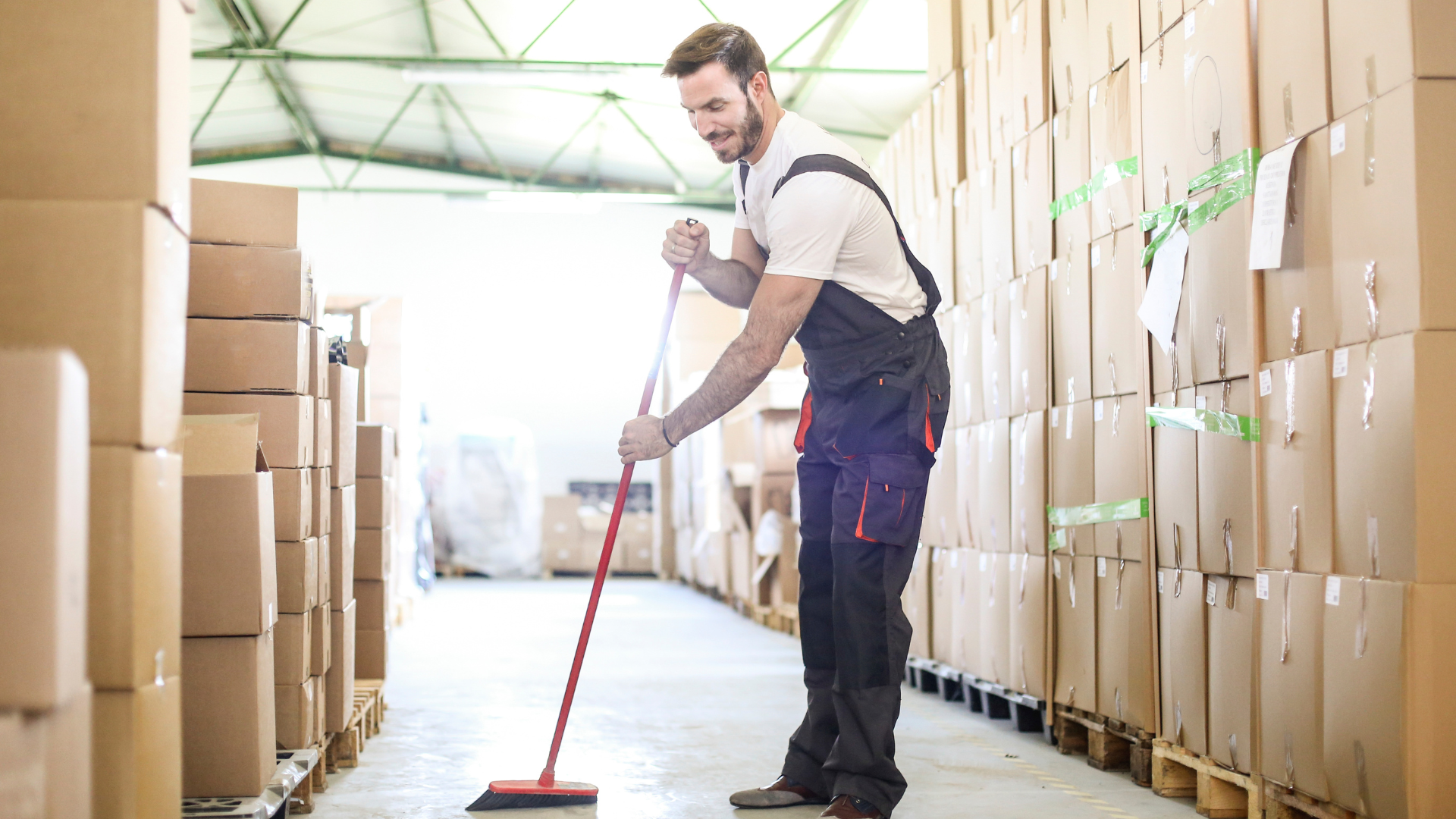 Worker mopping a warehouse aisle lined with stacked cardboard boxes