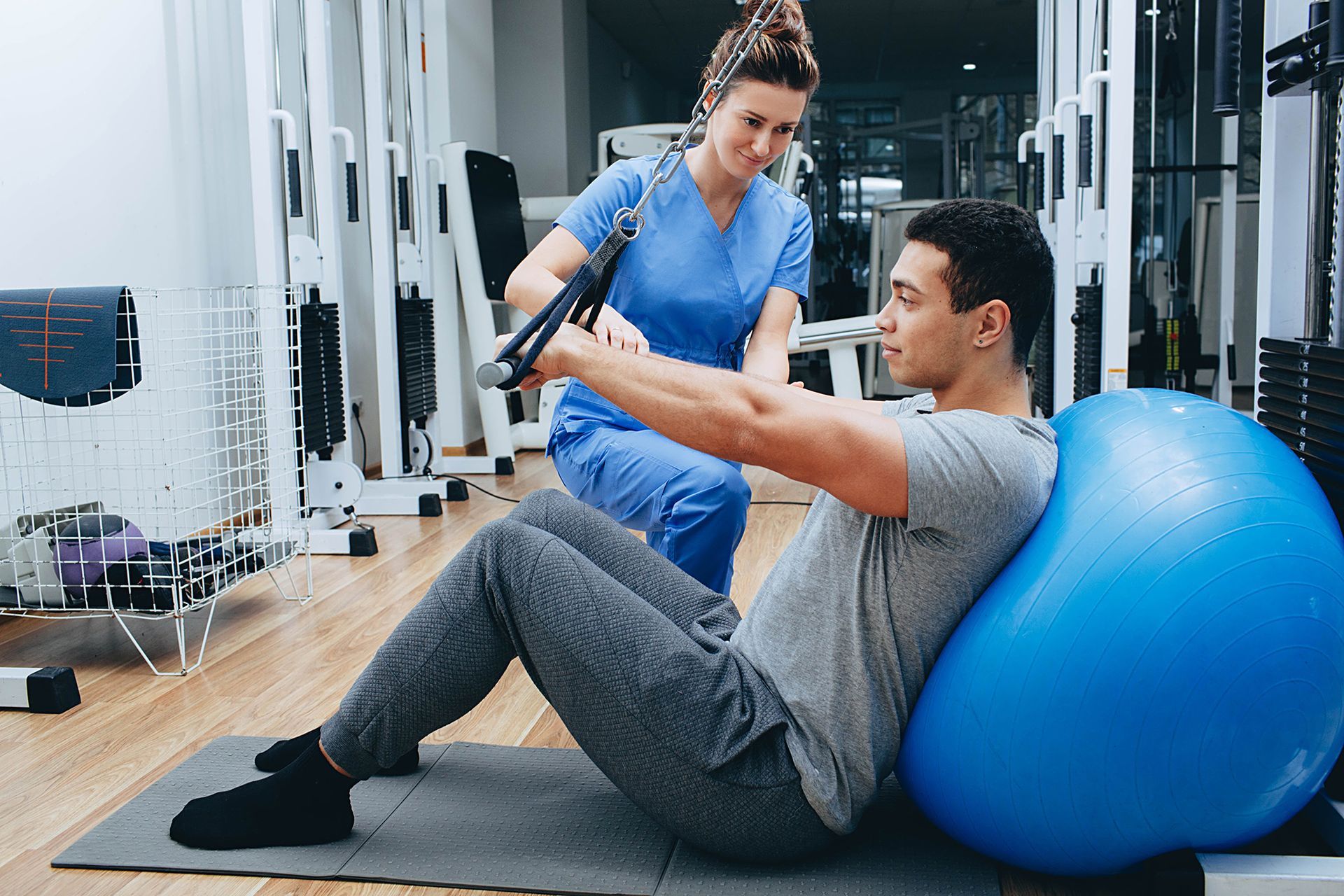 A man is doing exercises with a blue exercise ball while a nurse watches.