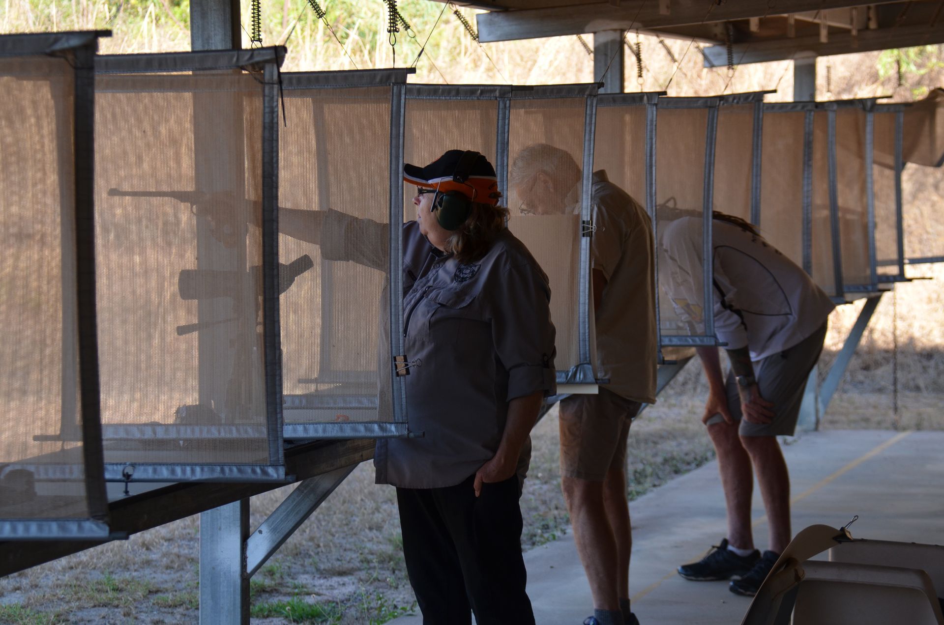 People at a shooting range, aiming rifles. They are behind mesh partitions in a sunny outdoor setting.