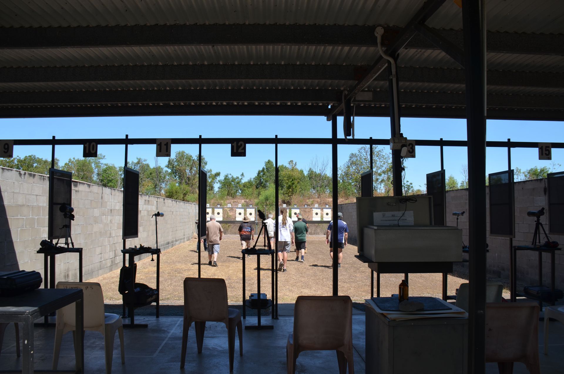 People at a shooting range, aiming at targets under a metal roof on a sunny day.