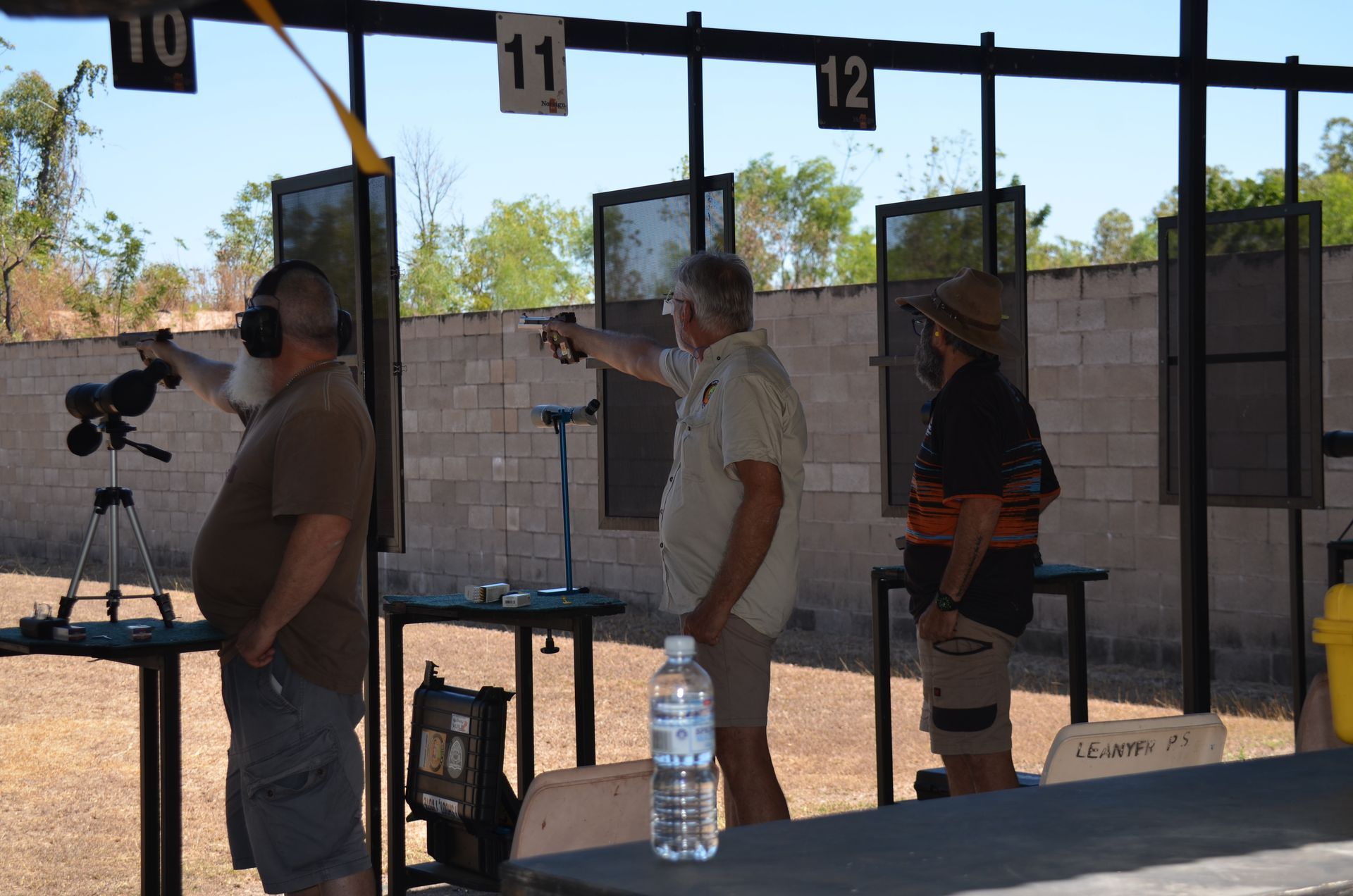 Three men at a shooting range. One aims a pistol, others observe. Bright sunlight.