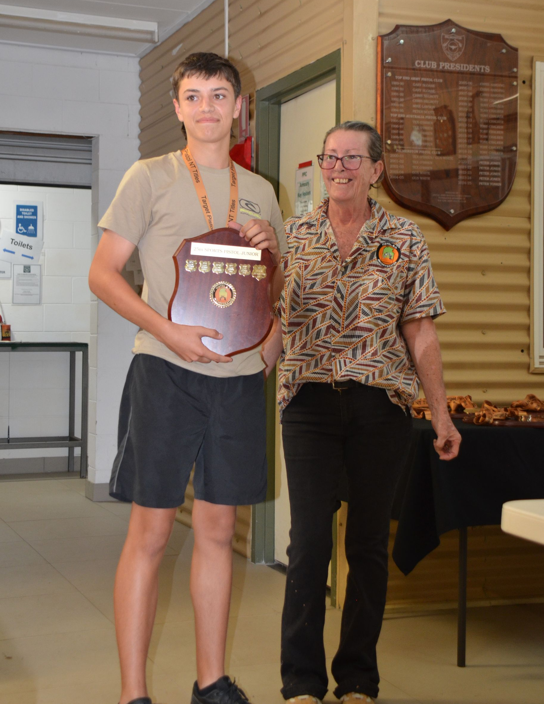 Teenager holding a trophy next to an older woman. Both smiling in a room with a wooden plaque on the wall.