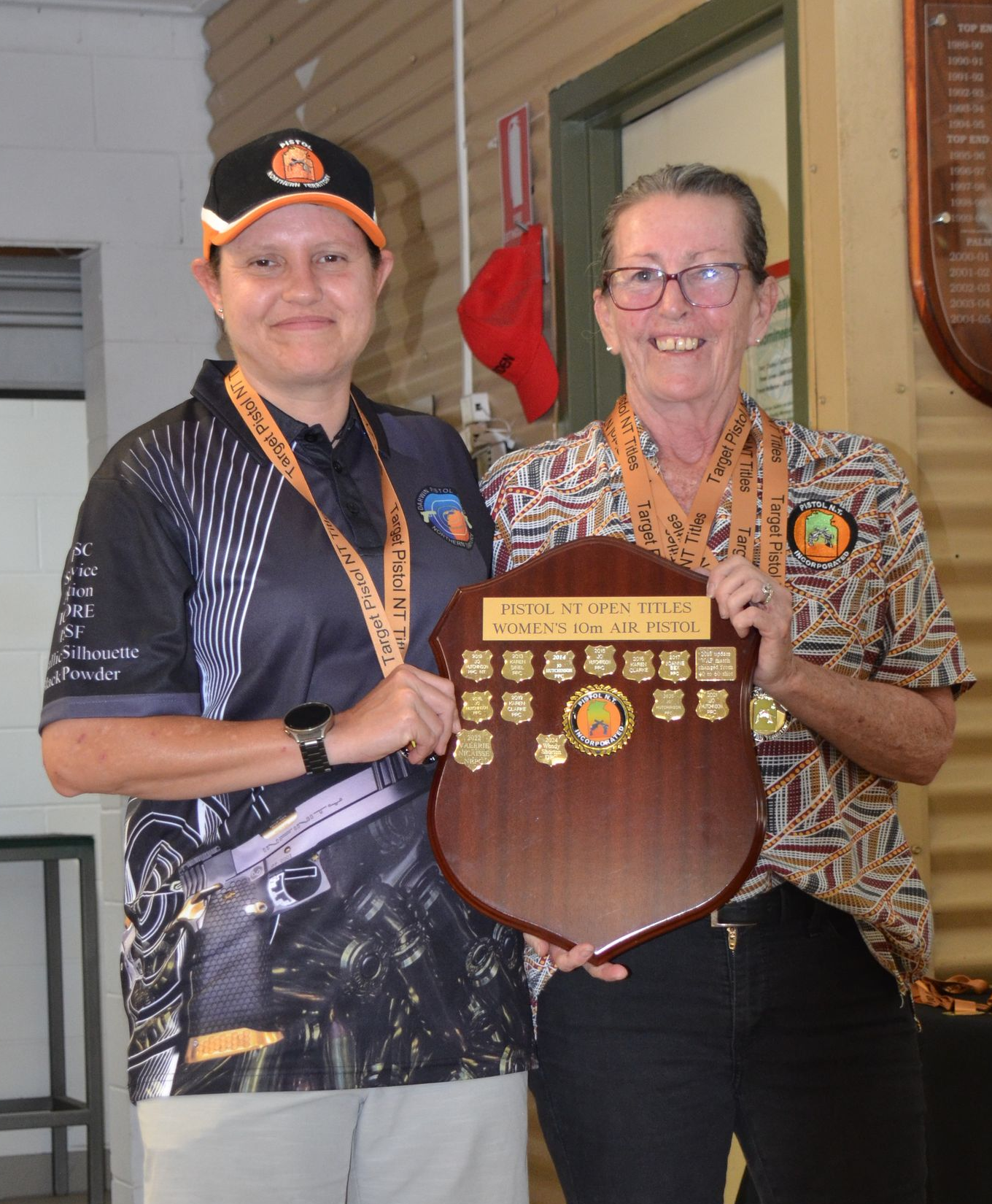 Two women holding a trophy, smiling, at an award ceremony. One wears a black shirt, the other a patterned shirt.