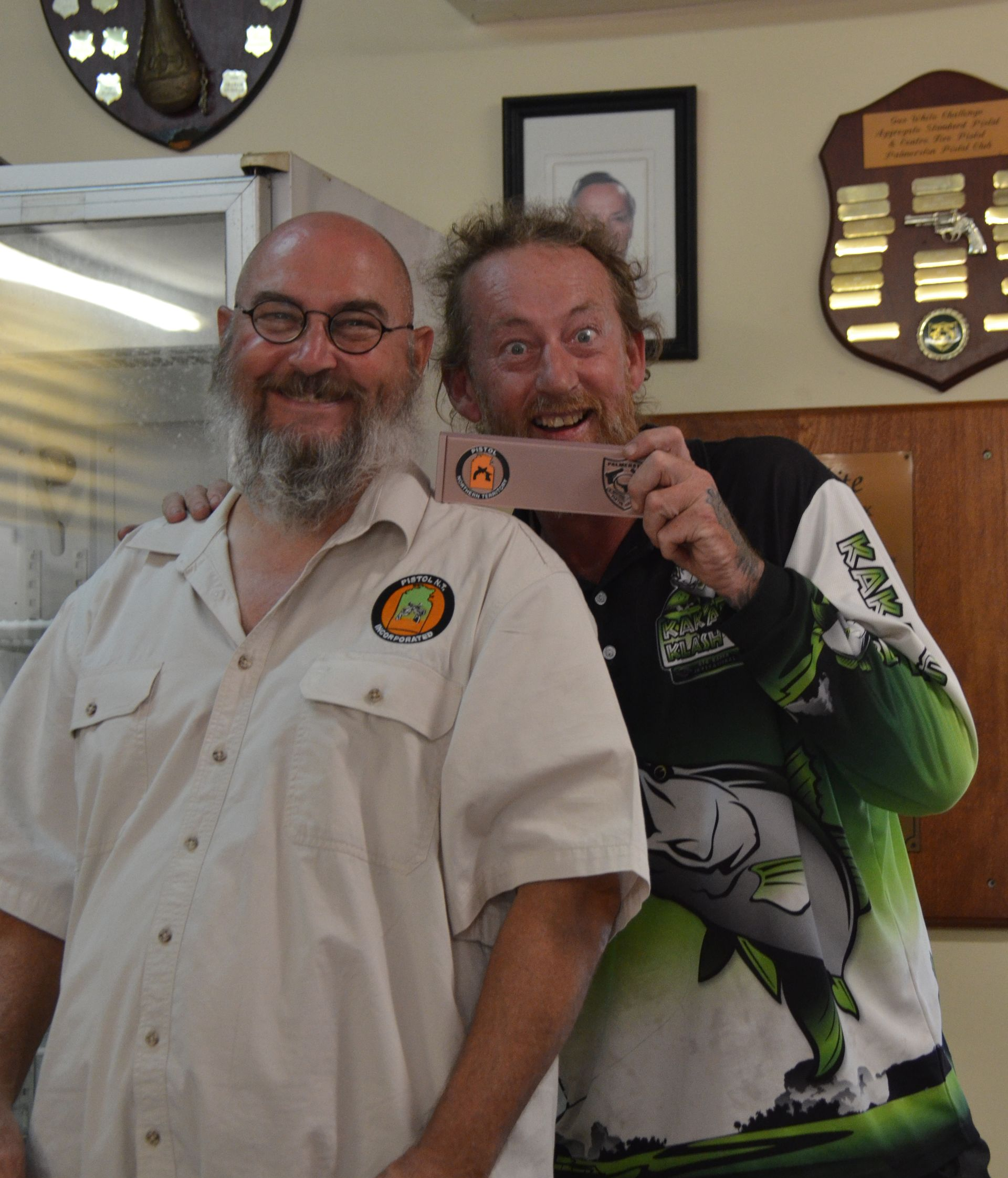 Two men smiling, one holding a trophy. Inside, awards on wall.