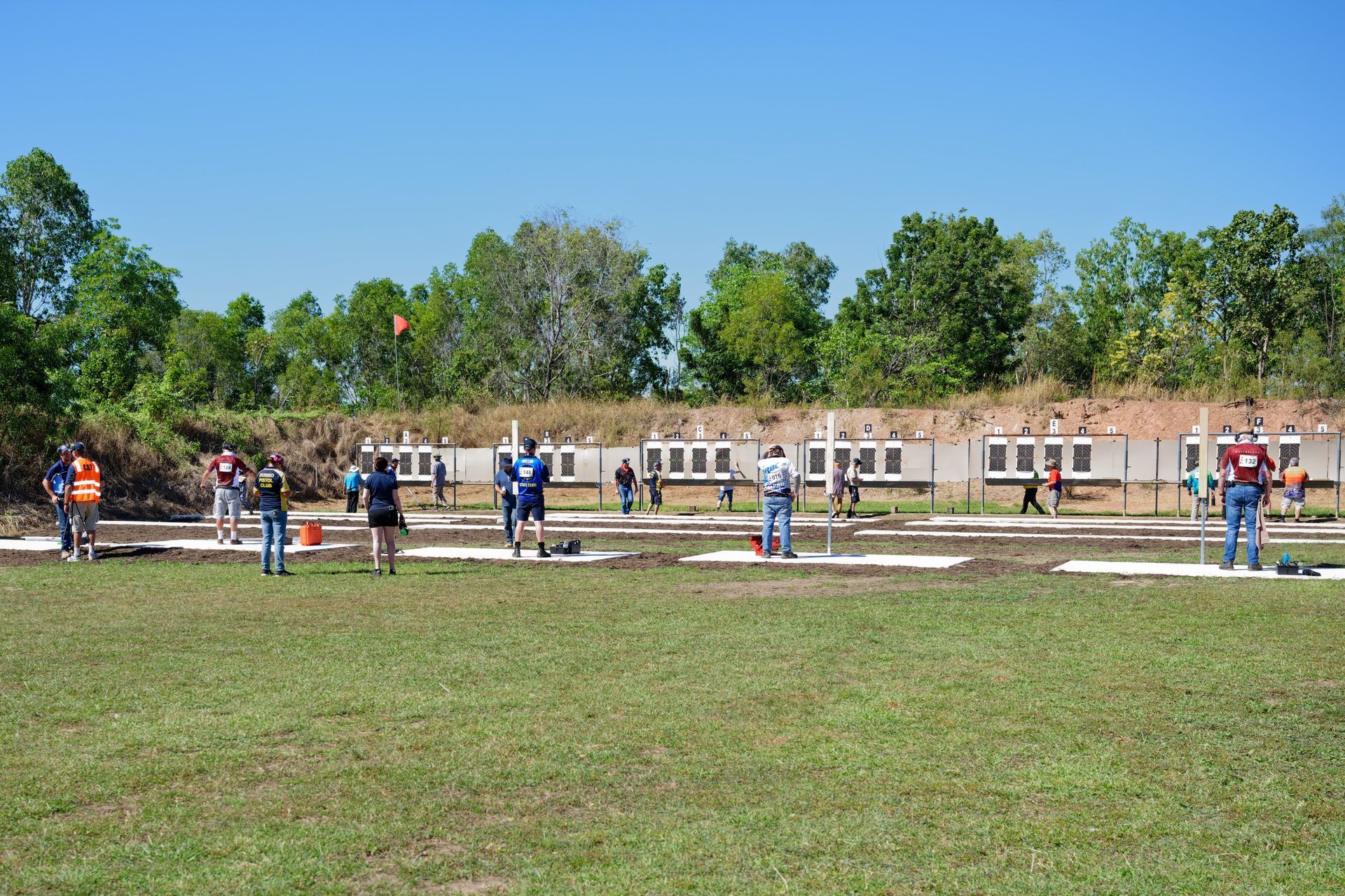 People shooting at targets on a sunny outdoor shooting range with trees in the background.