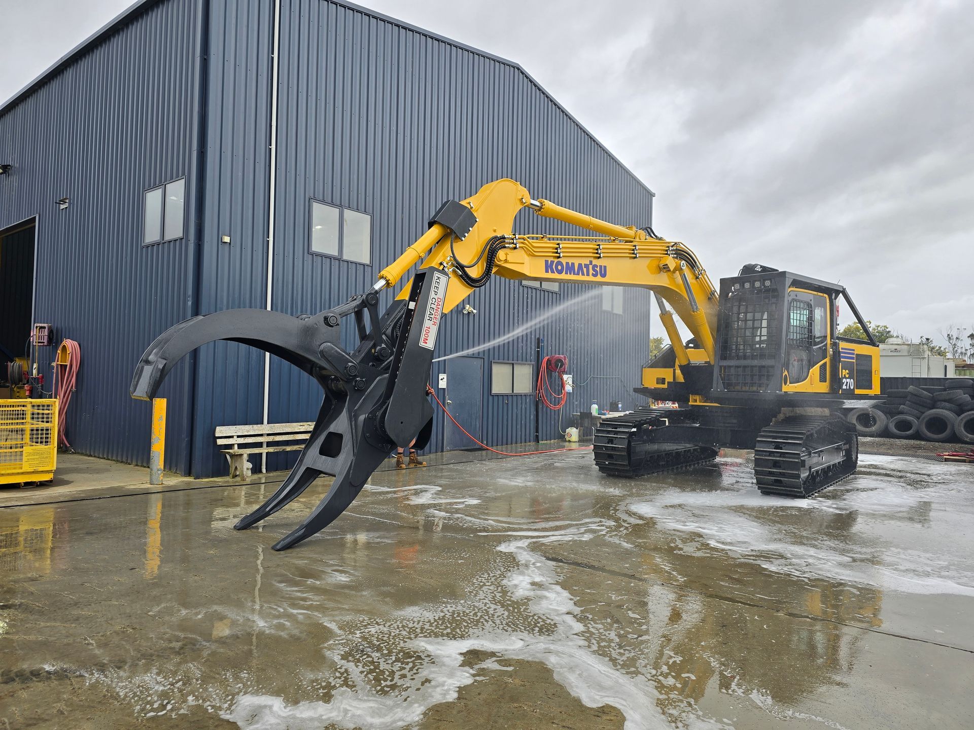 Yellow Komatsu excavator with grapple attachment being washed near a blue building.