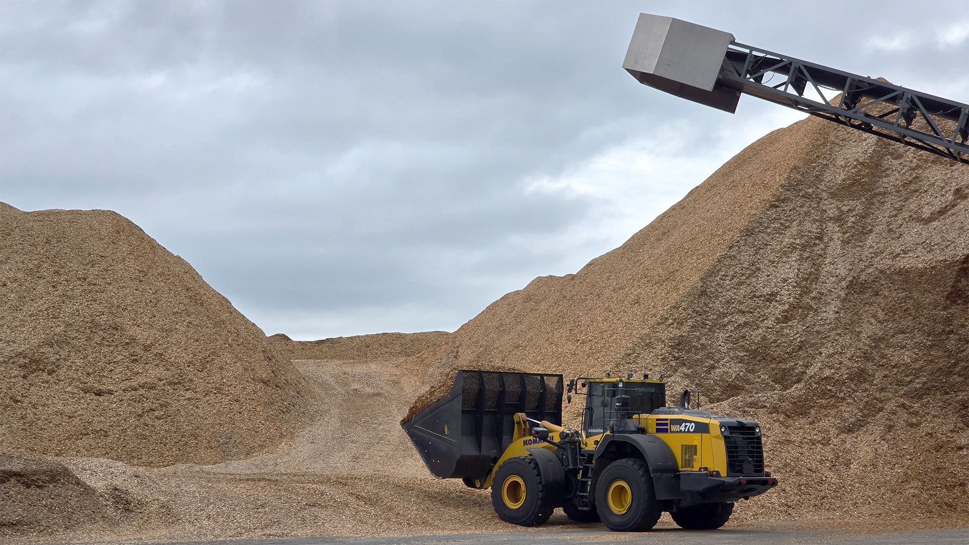 Yellow wheel loader in a gravel yard beneath large piles of wood chips, loading material from a conveyor.