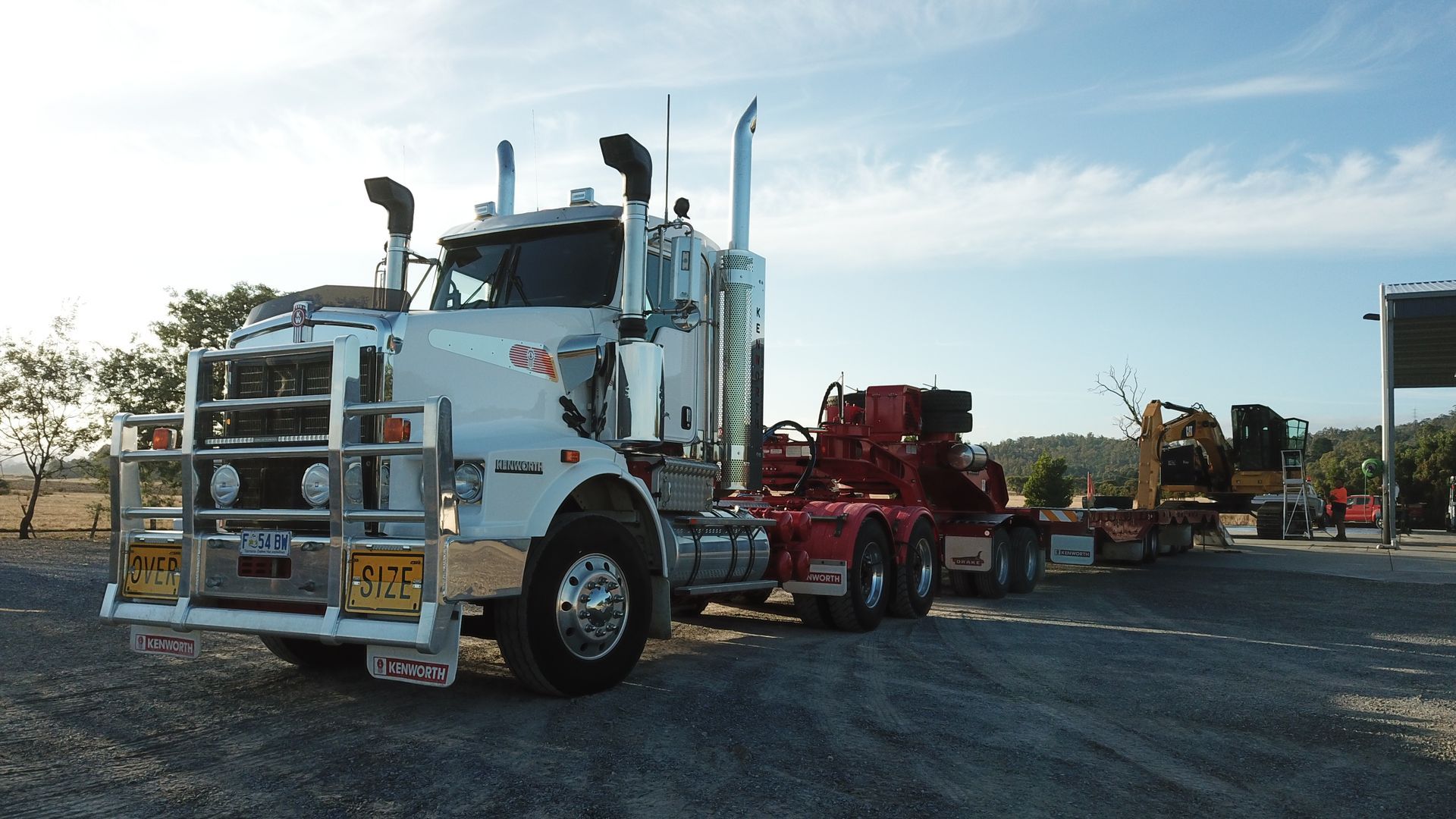 White semi-truck with equipment trailer parked on gravel, under a blue sky.