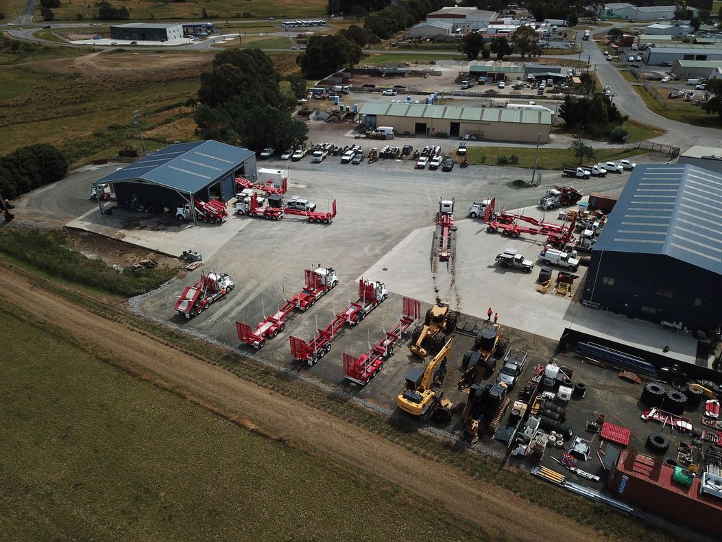 Aerial view of a heavy machinery yard with buildings, trucks, and equipment, on a rural setting with green fields.