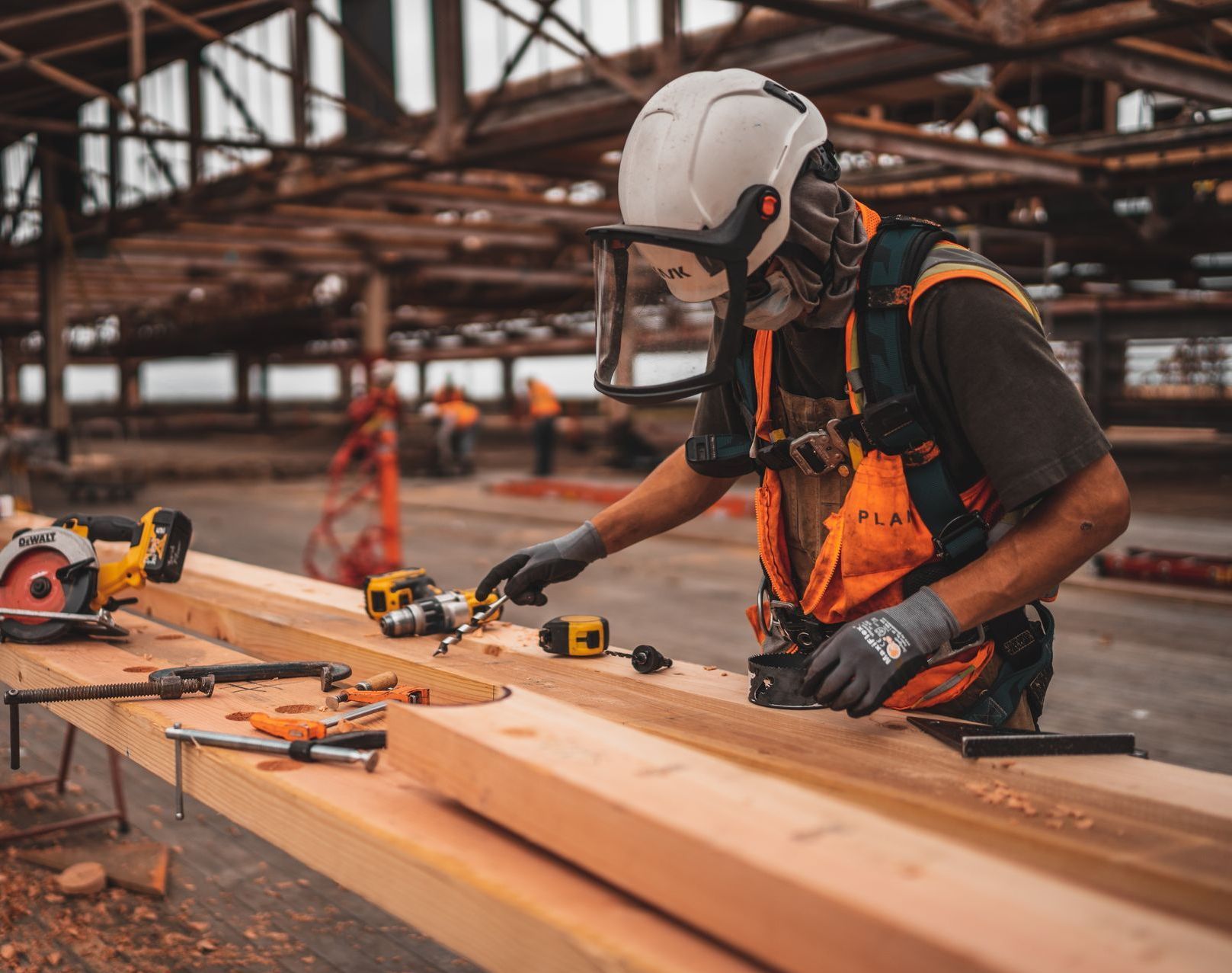 A man wearing a hard hat and safety vest is working on a wooden beam.